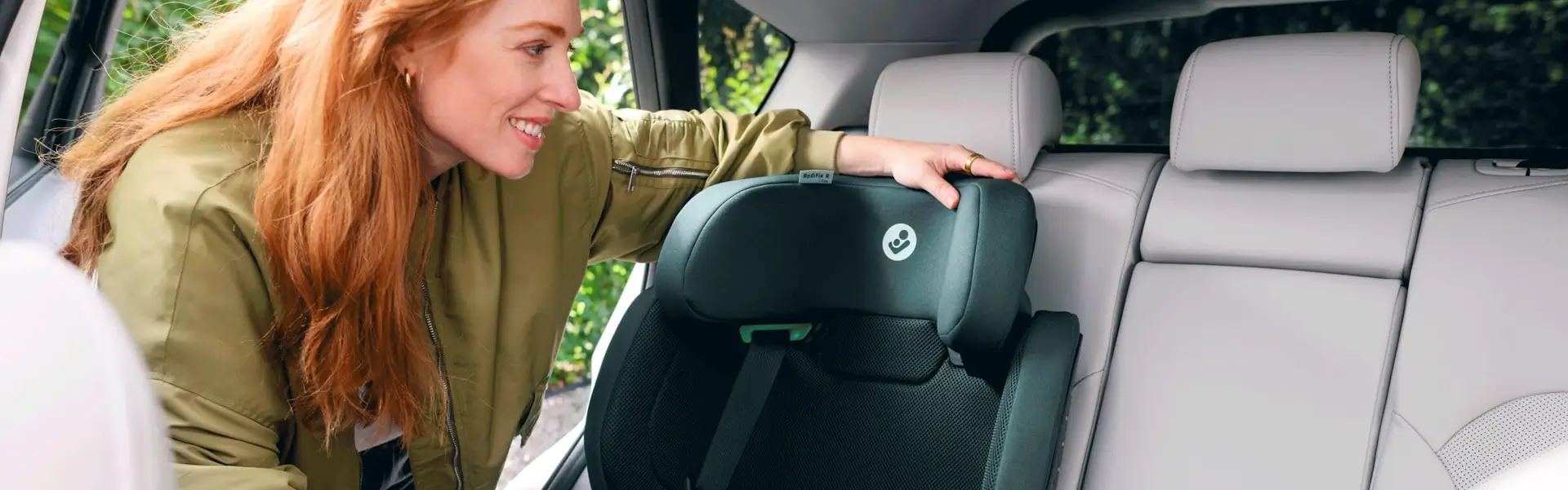Eine Frau lächelt, während sie den Maxi-Cosi RodiFix R i-Size-Kindersitz im Fond eines hellgrauen, sicher geparkten Autos einstellt.