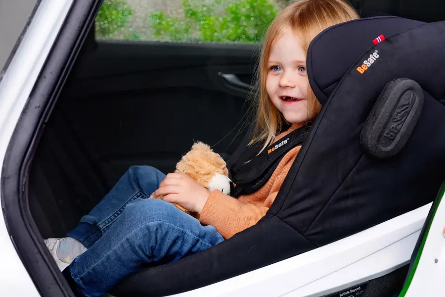 Ein Kind sitzt sicher in einem BeSafe Beyond-Autositz, hält ein Stofftier und lächelt, während es aus dem Fenster blickt.