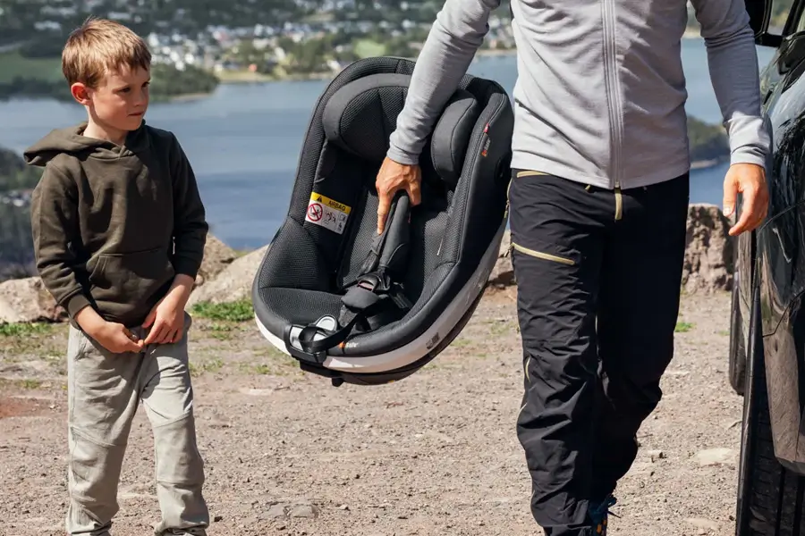 Vor einer malerischen Kulisse nähert sich ein Erwachsener, der den BeSafe Beyond-Autositz trägt, dem Fahrzeug, während ein kleiner Junge in der Nähe steht.