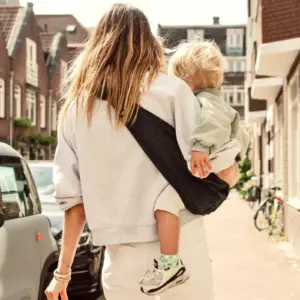 Eine Frau trägt mühelos ein Kleinkind in einer schwarzen Wildride Kindertrage aus Cord beim Spazierengehen.