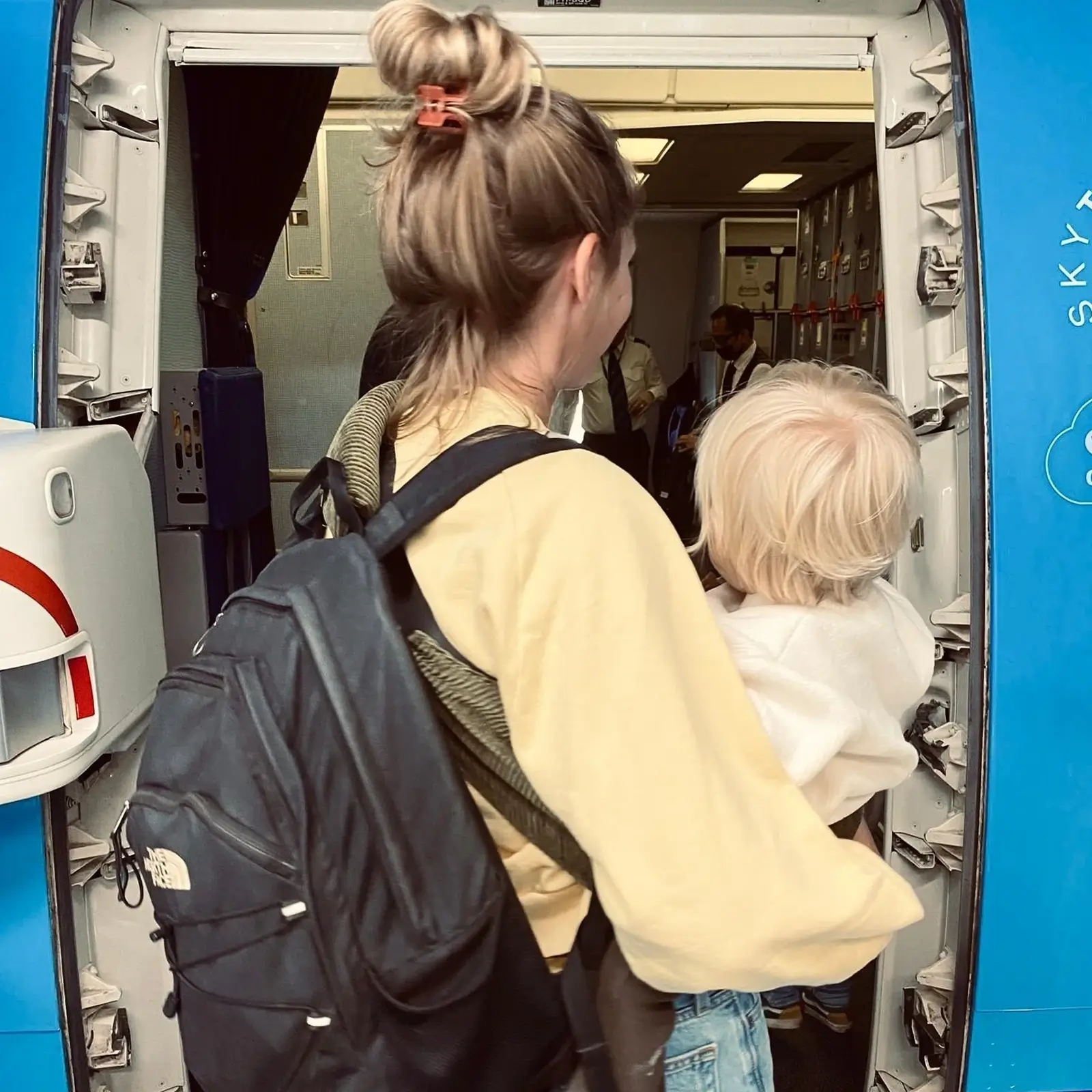 Eine Frau mit einer Wildride Kindertrage in Olivgrün trägt ein Kind in ein Flugzeug.