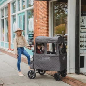 Eine Frau steht neben einem Kind, das in einem grauen Wonderfold-Wagen sitzt und durch die warme Abdeckung für W2 2.0 von Wonderfold Cold Weather Shield geschützt ist.